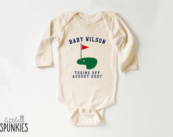 May include: A cream-colored long-sleeve baby bodysuit featuring a graphic of a green golf putting area with a red flag. The text reads BABY WILSON above the graphic and TEEING OFF AUGUST 2027 below the graphic in dark blue lettering.