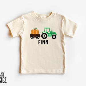 May include: A cream-colored short-sleeve t-shirt featuring a graphic of a green tractor pulling a brown wagon with an orange pumpkin. The name FINN is printed in black capital letters below the tractor graphic on the front of the shirt.