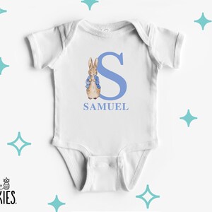 May include: A white short-sleeved baby bodysuit featuring a large light blue letter S, a watercolor illustration of a brown rabbit wearing a blue jacket, and the name SAMUEL printed in light blue capital letters below the letter and rabbit.