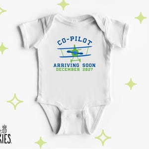 May include: A white short-sleeved baby bodysuit featuring a graphic of a blue and green airplane. The text reads CO-PILOT ARRIVING SOON DECEMBER 2027 in blue and green lettering. The design is centered on the chest of the garment.