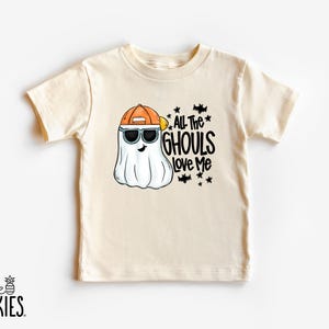May include: A cream-colored short-sleeve t-shirt featuring a graphic of a white ghost wearing an orange baseball cap and black sunglasses. The text "ALL THE GHOULS LOVE ME" is printed in black next to the ghost, accompanied by small bat and star icons.
