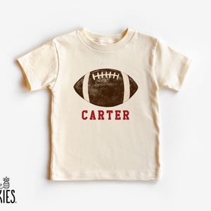 May include: Cream-colored t-shirt featuring a brown football graphic with white stripes and the name "CARTER" in maroon. The shirt has short sleeves and a crew neck. The brand logo "little SPUNKIES" is at the bottom left.