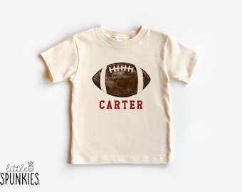 May include: A cream-coloured t-shirt with a brown American football graphic and white stripes, and the name "CARTER" in maroon. The shirt has short sleeves and a crew neck. The brand logo "little SPUNKIES" is at the bottom left.