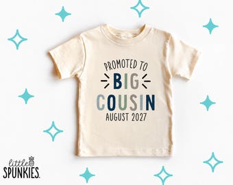 May include: A cream-colored short-sleeve t-shirt featuring the text PROMOTED TO BIG COUSIN AUGUST 2027 in navy, light blue, and grey lettering. The design includes small graphic starburst accents surrounding the word BIG.
