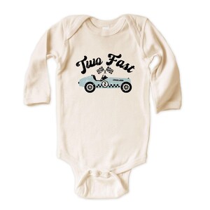 May include: A long-sleeve cream-colored baby bodysuit featuring a graphic of a light blue vintage race car with the number two on the side, two checkered flags, and the black text Two Fast printed in a distressed font above the car illustration.