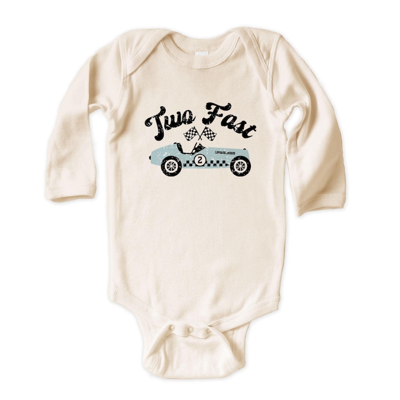 May include: A long-sleeve cream-colored baby bodysuit featuring a graphic of a light blue vintage race car with the number two on the side, two checkered flags, and the black text Two Fast printed in a distressed font above the car illustration.