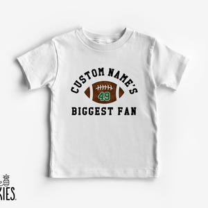May include: White t-shirt with the text "CUSTOM NAME'S BIGGEST FAN" in black and a brown football graphic with the number 49 in green. The shirt has short sleeves and a crew neck.