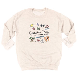 K&ouml;nnte beinhalten: Ein hellbeige Sweatshirt mit langen &Auml;rmeln. Auf der Vorderseite befindet sich eine farbenfrohe Grafik mit dem Text "Cousin Crew Myrtle Beach 2025", umgeben von Strandmotiven wie Sonne, Palmen und Flip-Flops.
