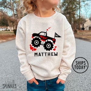 May include: A light beige sweatshirt featuring a black monster truck design with red accents and hearts. The name "MATTHEW" is printed below the truck. The sweatshirt has a crew neck and long sleeves.