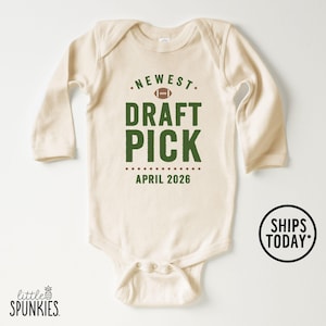 May include: Cream-colored baby onesie with long sleeves. The front features the text "NEWEST DRAFT PICK" in green, with a football graphic above and "APRIL 2026" below. A small logo and the words "SHIPS TODAY" are also visible.