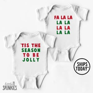 May include: Two white baby onesies with holiday-themed text. One reads "'TIS THE SEASON TO BE JOLLY" in red and green. The other says "FA LA LA LA LA" in the same colors. The brand logo "little SPUNKIES" is visible.