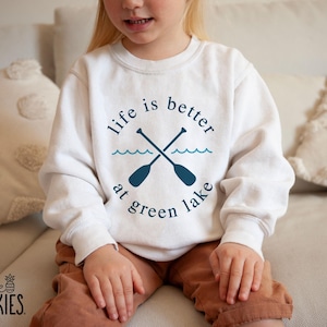 May include: A white sweatshirt with the text "life is better at green lake" in a teal blue color. The design includes crossed oars and wavy lines. The sweatshirt is worn by a child.