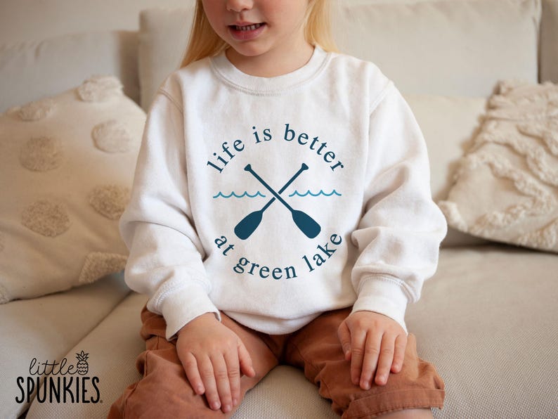 May include: A white sweatshirt with the text "life is better at green lake" in a teal blue color. The design includes crossed oars and wavy lines. The sweatshirt is worn by a child.