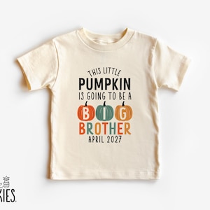 May include: A cream-colored short-sleeve t-shirt featuring the black text THIS LITTLE PUMPKIN IS GOING TO BE A BIG BROTHER APRIL 2027. The word BIG is displayed with three pumpkin illustrations in orange, teal, and orange, each containing a white letter.