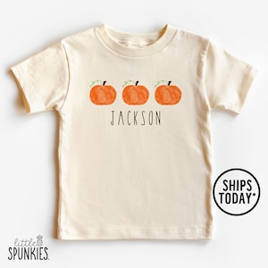 May include: Cream-colored short-sleeved t-shirt featuring three orange pumpkin graphics with green vines. The name "JACKSON" is printed in black below the pumpkins. A round sticker with "SHIPS TODAY" is on the right.
