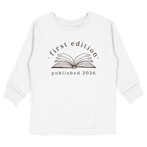 May include: A white long-sleeve shirt featuring a dark brown graphic of an open book. Above the book, the text reads "first edition." and below the book, the text reads "published 2026". The design is centered on the chest of the shirt.