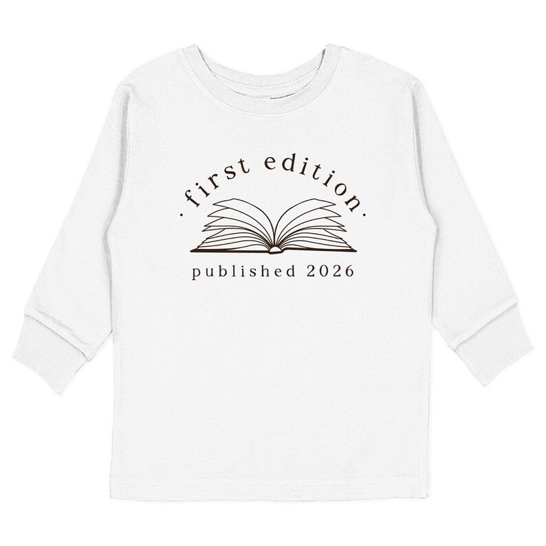 May include: A white long-sleeve shirt featuring a dark brown graphic of an open book. Above the book, the text reads "first edition." and below the book, the text reads "published 2026". The design is centered on the chest of the shirt.