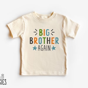 May include: A cream-colored short-sleeve t-shirt featuring the text BIG BROTHER AGAIN in a multi-colored font. The word BIG is in green, BROTHER is in dark gray and orange, and AGAIN is in black, flanked by two light blue stars and black dash accents.