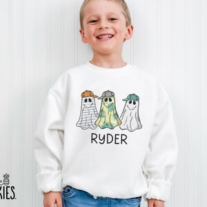 May include: A white long-sleeve sweatshirt featuring a graphic of three ghosts wearing baseball caps in brown, gray, and green. The ghosts have different patterns including stripes and spots. The name RYDER is printed in black text below the ghosts.