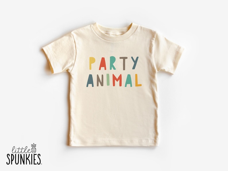 May include: A cream-colored short-sleeve t-shirt featuring the text PARTY ANIMAL printed in a multi-colored, sans-serif font. The letters are colored in yellow, red, teal, brown, and blue, arranged in two lines across the center of the chest.
