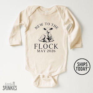 May include: Cream-colored baby onesie with long sleeves. The front features a black graphic of a lamb and the text "NEW TO THE FLOCK MAY 2026". The onesie has a round neckline and snap closures.