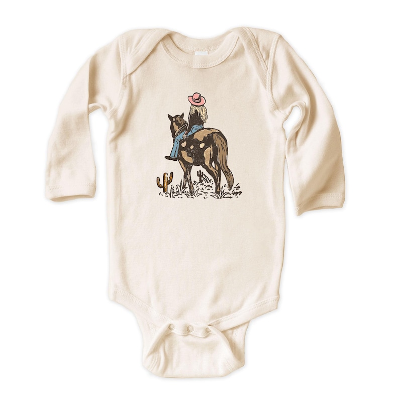 Puede incluir: Body de beb&eacute; color crema con mangas largas. El body presenta un gr&aacute;fico de una persona montando a caballo, con un sombrero rosa y vaqueros azules. Un cactus tambi&eacute;n est&aacute; en el gr&aacute;fico.