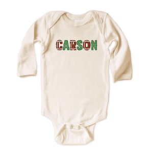 Op de afbeelding: Cr&egrave;mekleurige baby-onesie met lange mouwen. De naam "CARSON" staat in groene en voetbal-textuur letters op de borst. De onesie heeft drukknoopsluitingen aan de onderkant.