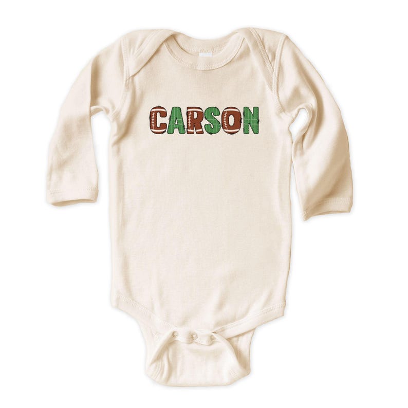 Op de afbeelding: Cr&egrave;mekleurige baby-onesie met lange mouwen. De naam "CARSON" staat in groene en voetbal-textuur letters op de borst. De onesie heeft drukknoopsluitingen aan de onderkant.
