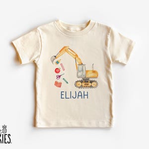 Puede incluir: Camiseta de manga corta color crema con una ilustraci&oacute;n en acuarela de una excavadora amarilla con &uacute;tiles escolares. El nombre "ELIJAH" est&aacute; impreso en azul marino debajo del dise&ntilde;o. El logotipo de la marca "little SPUNKIES" est&aacute; en la parte inferior izquierda.