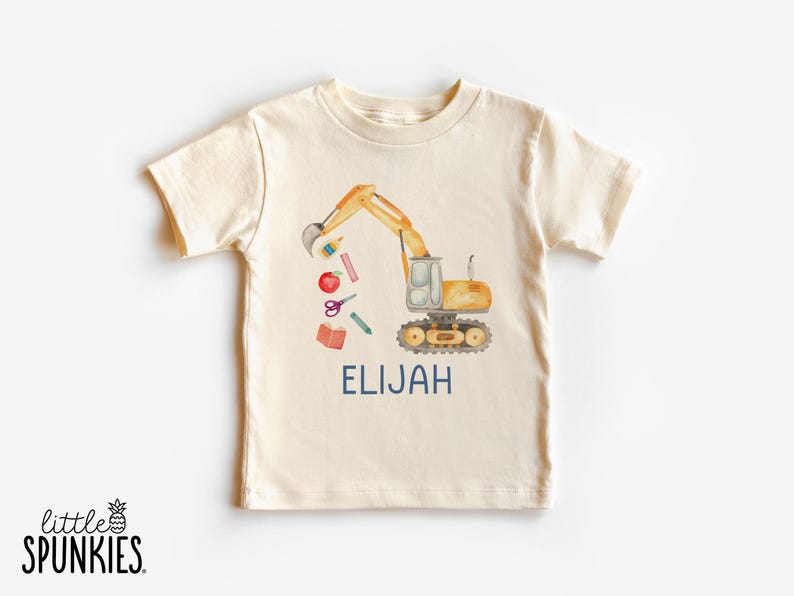 Puede incluir: Camiseta de manga corta color crema con una ilustraci&oacute;n en acuarela de una excavadora amarilla con &uacute;tiles escolares. El nombre "ELIJAH" est&aacute; impreso en azul marino debajo del dise&ntilde;o. El logotipo de la marca "little SPUNKIES" est&aacute; en la parte inferior izquierda.