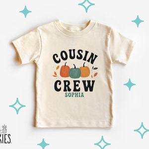 May include: A cream-colored short-sleeve t-shirt featuring the black text COUSIN CREW above three pumpkins in orange, teal, and orange. Below the pumpkins, the name SOPHIA is printed in teal. The design includes small scattered autumn leaves.