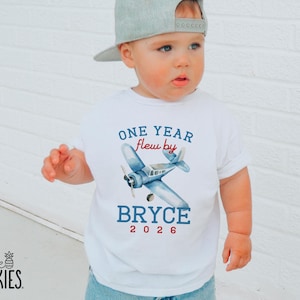May include: A white short-sleeved t-shirt featuring a blue watercolor airplane graphic. The text reads ONE YEAR flew by BRYCE 2026 in blue and red lettering. The shirt is paired with a light gray baseball cap, creating a simple and clean look.