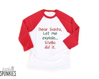 May include: A white raglan shirt with red three-quarter length sleeves. The front features the text Dear Santa, Let me explain... Stella did it. The words Dear, Santa, Stella, and did it are in red, while the words Let me explain are in green.