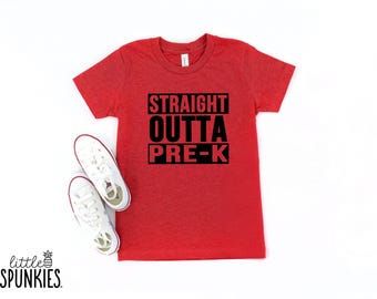 May include: A red t-shirt with the text "STRAIGHT OUTTA PRE-K" in black block letters. White canvas sneakers with red trim are next to the shirt. The shirt is a youth size.