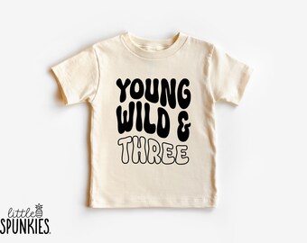 May include: A cream-colored short-sleeve t-shirt featuring the black text "YOUNG WILD & THREE" printed on the front. The words "YOUNG" and "WILD" are in a solid black font, while the word "THREE" is in a black outlined font style.
