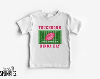 May include: A white short-sleeve t-shirt featuring a graphic of a green football field with a pink football in the center. The text "TOUCHDOWN" appears in pink above the field, and the text "KINDA DAY" appears in pink below the field.