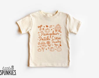 May include: A cream-colored short-sleeve t-shirt featuring a burnt orange graphic print. The design includes the text "Pumpkin Patch Crew" and "Paisley" surrounded by illustrations of pumpkins, a scarecrow, leaves, and a wagon.
