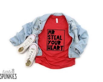 May include: A red t-shirt featuring the black text MR STEAL YOUR HEART inside a black rectangular border, layered under a light blue denim jacket, accompanied by a pair of white canvas trainers with white laces and rubber soles.