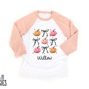 May include: A white raglan shirt with peach-colored sleeves featuring a graphic print of orange and pink jack-o'-lantern pumpkins interspersed with black bows and small stars. The name Willow is printed in black script lettering below the design.