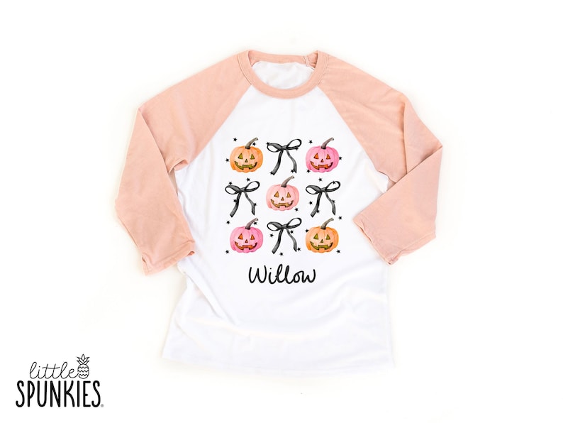 May include: A white raglan shirt with peach-colored sleeves featuring a graphic print of orange and pink jack-o'-lantern pumpkins interspersed with black bows and small stars. The name Willow is printed in black script lettering below the design.