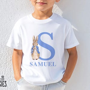 May include: A white short-sleeved t-shirt featuring a large light blue letter S, the name SAMUEL in light blue capital letters, and a watercolor-style illustration of a brown rabbit wearing a blue jacket standing next to the letter.