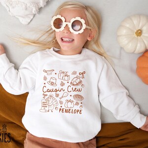 May include: A white long-sleeve sweatshirt featuring a brown graphic print of Thanksgiving-themed icons including a turkey, pumpkins, a pie, and a gravy boat. The text reads Cousin Crew and PENELOPE in a brown font, with small leaf accents.