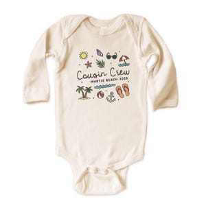 K&ouml;nnte beinhalten: Cremefarbener Baby-Body mit langen &Auml;rmeln. Auf der Vorderseite befindet sich eine farbenfrohe Grafik mit dem Text "Cousin Crew Myrtle Beach 2025" und Strand-Illustrationen, darunter eine Sonne, Sonnenbrille und Flip-Flops.