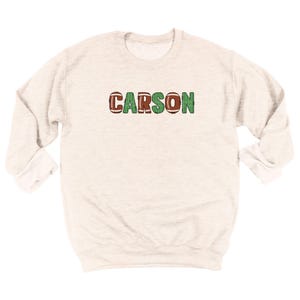 Op de afbeelding: Een lichtgrijs sweatshirt met de naam "CARSON" over de borst. De letters "C", "A", "R" en "N" zijn ontworpen als American footballs, terwijl de letters "O" en "S" groen zijn.