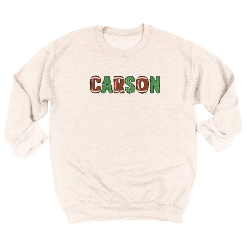 Op de afbeelding: Een lichtgrijs sweatshirt met de naam "CARSON" over de borst. De letters "C", "A", "R" en "N" zijn ontworpen als American footballs, terwijl de letters "O" en "S" groen zijn.