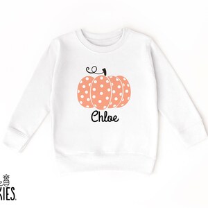 May include: A white long-sleeve sweatshirt featuring a central graphic of a peach-colored pumpkin with white polka dots and a black stem. The name Chloe is printed in black script lettering directly below the pumpkin graphic on the front.
