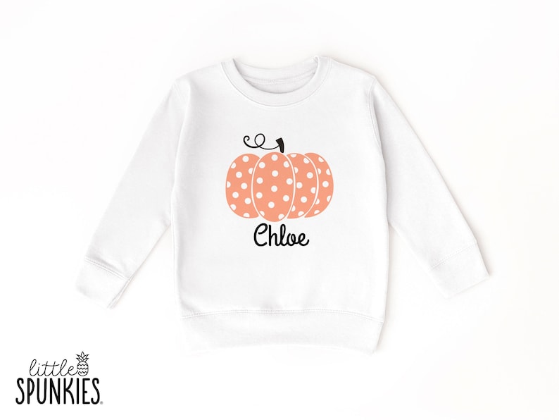 May include: A white long-sleeve sweatshirt featuring a central graphic of a peach-colored pumpkin with white polka dots and a black stem. The name Chloe is printed in black script lettering directly below the pumpkin graphic on the front.