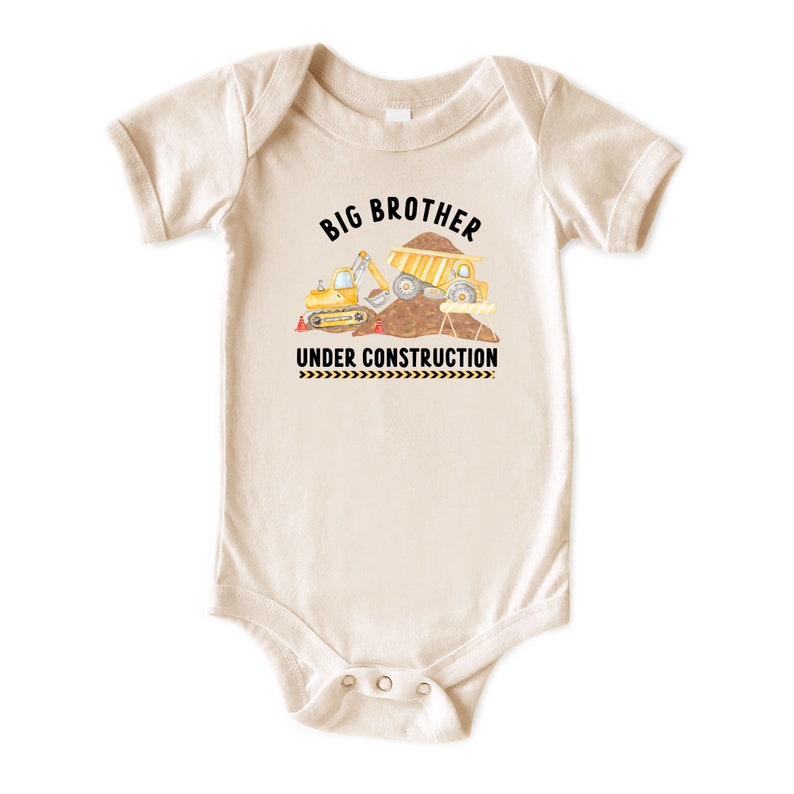 May include: A beige short-sleeve bodysuit featuring a graphic of a yellow excavator and dump truck on a dirt pile. The text reads BIG BROTHER above the vehicles and UNDER CONSTRUCTION below, with a black and yellow hazard stripe pattern at the bottom.