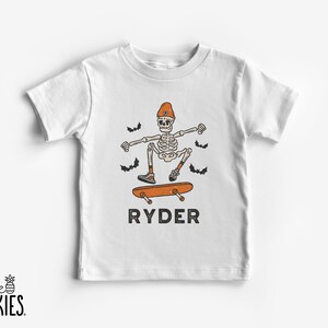 May include: A white short-sleeve t-shirt featuring a graphic of a skeleton wearing an orange beanie and sneakers while riding an orange skateboard. The design includes four small black bats and the name RYDER printed in black capital letters.