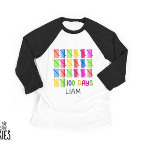 May include: White baseball style shirt with black sleeves. The shirt features colorful vertical lines in a grid pattern, with the text "100 DAYS" and the name "LIAM" in colorful letters. The brand logo "little SPUNKIES" is at the bottom.